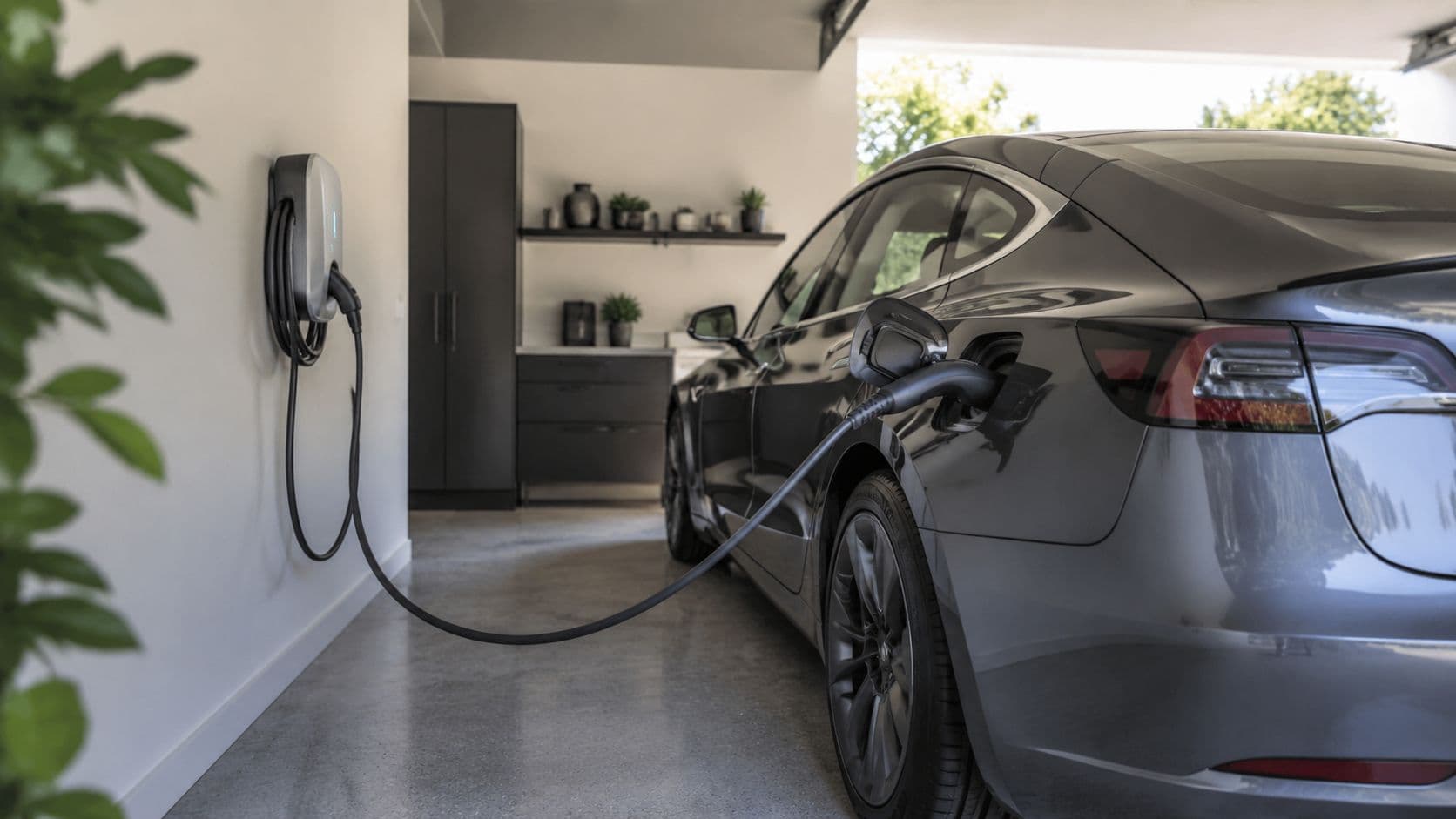 Un coche eléctrico de color gris cargando su batería mediante un cargador de pared en un garaje.